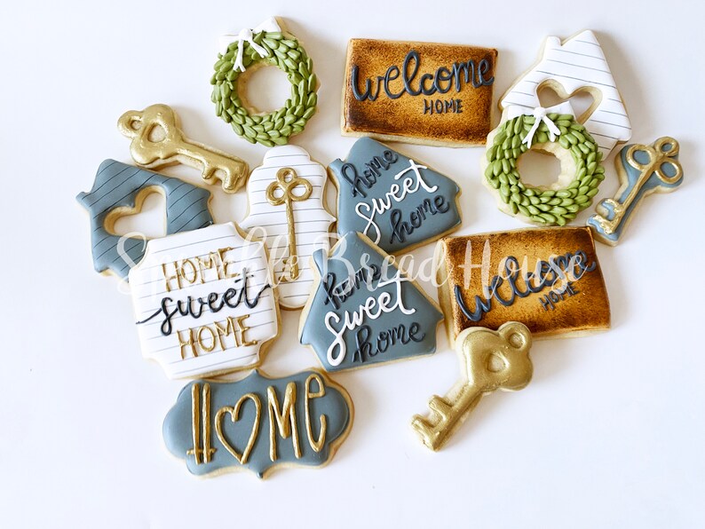Puede incluir: Un surtido de galletas decoradas con tem&aacute;tica de hogar. Las galletas incluyen formas de casa, llaves, coronas y letreros con frases como "Welcome Home" y "Home Sweet Home" en glaseado dorado y negro. Las galletas est&aacute;n sobre una superficie blanca.