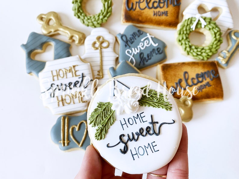 Puede incluir: Surtido de galletas de az&uacute;car decoradas con tem&aacute;tica de hogar. Las galletas incluyen formas de llaves, casas y coronas, con las frases "Home Sweet Home" y "Welcome Home" escritas en elegante caligraf&iacute;a. Los colores incluyen blanco, dorado, verde y gris.