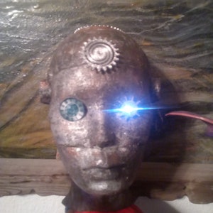 May include: A silver robot head with a red, white, and blue shirt. The robot has one blue eye and one green eye. The robot has a silver gear on its forehead.