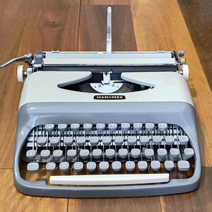May include: A classic Hanimex typewriter in grey and cream, with a full QWERTY keyboard layout. The typewriter has a silver carriage return lever and a paper guide. The brand name "HANIMEX" is displayed on the front.