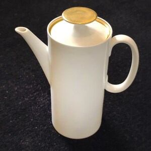 May include: White ceramic coffee pot with a gold lid and handle. The pot has a cylindrical shape and a simple design.