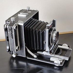 May include: A black and silver large format camera with a bellows system and a lens with a depth of field scale. The camera is on a black surface.