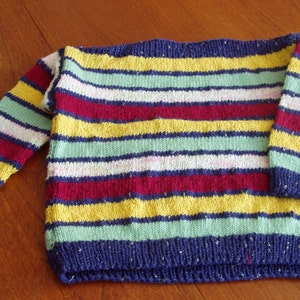 Könnte beinhalten: Ein gestreifter Strickpullover in den Farben Blau, Grün, Gelb, Rot und Weiß. Der Pullover hat einen weiten Ausschnitt und lange Ärmel.