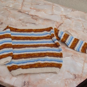 Könnte beinhalten: Ein gestreifter Pullover mit braunen, weißen und blauen Streifen. Der Pullover ist gestrickt und hat eine lockere Passform.