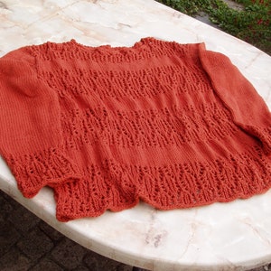 Puede incluir: Un suéter de punto de color óxido con un patrón de encaje. El suéter tiene un cuello redondo y mangas largas.