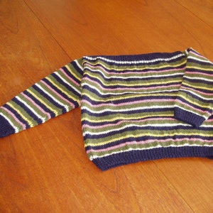 Könnte beinhalten: Ein gestreifter Strickpullover in Marineblau, Weiß, Olivgrün, Rosa und Gelb. Der Pullover hat lange Ärmel und einen marineblauen Rippsaum und Bündchen. Das Kleidungsstück wird auf einer Holzoberfläche präsentiert.