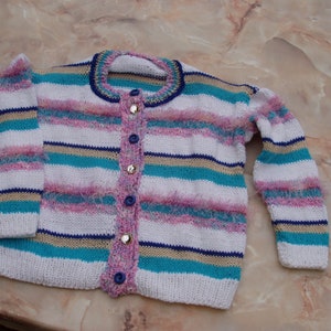Könnte beinhalten: Ein gestreifter Strickcardigan mit Knopfleiste. Der Cardigan ist weiß mit blauen, rosa und gelben Streifen. Der Cardigan hat eine flauschige Textur.