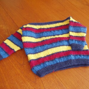 Könnte beinhalten: Ein gestreifter Pullover in Blau, Gelb und Burgunderrot. Das Strickstück hat horizontale Farbstreifen und einen Rundhalsausschnitt. Der Pullover ist für ein Kind entworfen.