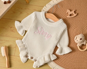 Può includere: Un body bianco in maglia per neonati, con maniche lunghe e dettagli a balze. Il body ha la parola "Sherry" ricamata in rosa sul davanti. Presenta bottoni in legno ed è esposto su una gruccia di legno.