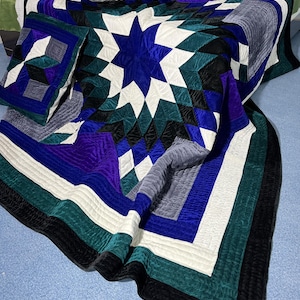 May include: A quilted bedspread and two matching pillows in shades of blue, purple, black, white, and teal. The bedspread features a large star design in the center. The pillows have geometric patterns. The quilt is made of a soft, textured fabric.