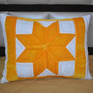 May include: A white pillow with a large yellow star design. The star is made of multiple pieces of fabric sewn together to create a quilted effect. The pillow is on a bed with a brown headboard.