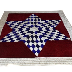 May include: A large, handmade quilt featuring a central star design. The star is composed of blue and white diamond shapes, set against a deep red background. The quilt is bordered with a white, quilted edge.
