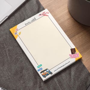 May include: A personalized notepad with a travel theme. The notepad features a cream-colored interior with a white border and yellow accents. Illustrations include a plane, tuk-tuk, passport, and travel-related icons. The name "Emily" is printed at the top.