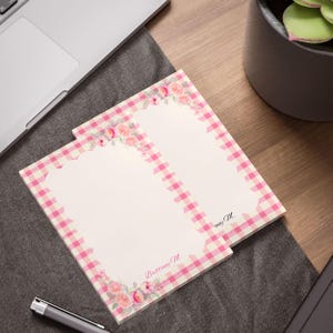 May include: Two floral-themed notepads with a pink gingham border and a floral arrangement at the top. The notepads have the name "Brittany M." printed on them. The notepads are on a wooden desk next to a laptop and a pen.