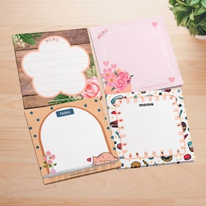 May include: Four square memo pads with different designs. One pad features a flower shape with lines, another has pink hearts and flowers, a third has a cat illustration, and the last has a food-themed border. Each pad has the word "memo" or "notes".