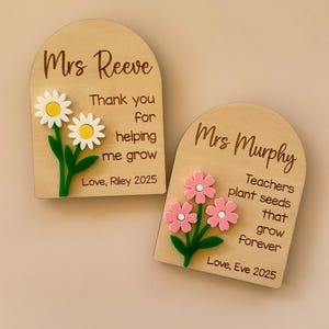 May include: Two wooden plaques with arched tops. One says "Mrs Reeve" with a daisy design and the text "Thank you for helping me grow." The other says "Mrs Murphy" with pink flowers and the text "Teachers plant seeds that grow forever."