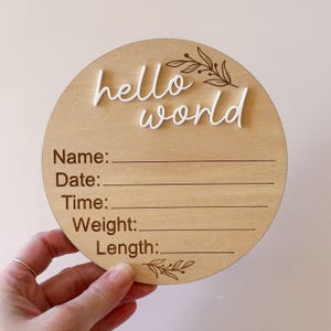 May include: A circular wooden sign with the words "hello world" in white script. The sign has lines for Name, Date, Time, Weight, and Length. Decorative floral elements are at the top and bottom. The sign is held by a hand.