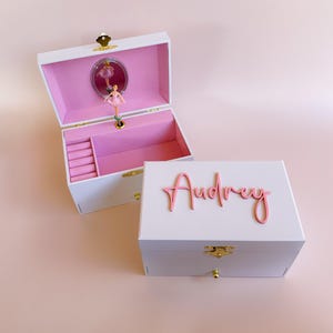 May include: A white jewellery box with a pink interior, featuring a rotating ballerina figurine in front of a small oval mirror. The lid is personalised with the name Audrey in pink script lettering, and the box includes a gold latch and drawer.