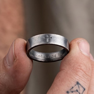 May include: A brushed dark grey metal band ring featuring a small engraved cross on the outer surface and the text Joshua 24:15 engraved on the inner band. The ring has a flat profile and a matte finish, showcasing a simple and clean design.