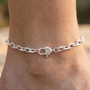 May include: A silver chain anklet with a heart-shaped charm. The anklet is made of small, oval-shaped links. The heart charm has the initials "LH" engraved on it. The anklet is worn on an ankle.