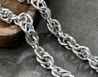 May include: Close-up of two silver-colored chain necklaces with a rope-like design. The chains are displayed on a wooden tray and a gray surface. The links are tightly woven, creating a textured appearance. The chains are likely jewelry.