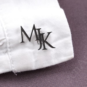 May include: Silver cufflinks with the initials "MK" displayed on a white shirt cuff. The cufflinks are in a serif font and are positioned on the cuff of a white dress shirt. The background is a textured, purple fabric.