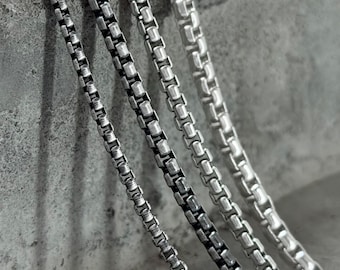May include: Close-up of several silver box chain necklaces. The chains are displayed against a textured gray background. The chains vary slightly in thickness and have a polished finish, reflecting light.
