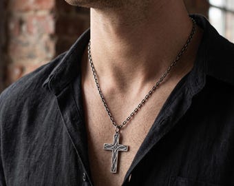 May include: A silver cross pendant necklace with an elaborate pattern. The cross is suspended from a matching chain, contrasting with the dark shirt. The necklace is a religious or fashion accessory.