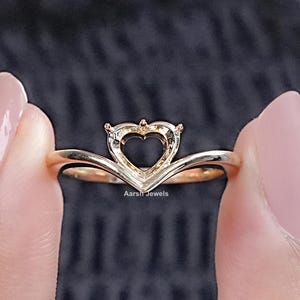 Op de afbeelding: Een gouden hartvormige ringsetting met een delicate, gebogen band. De setting is ontworpen om een ​​edelsteen in het midden te houden.