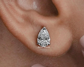 May include: A single pear-shaped diamond stud earring in a silver setting. The earring is worn in the left ear of a person with fair skin and pink nail polish.