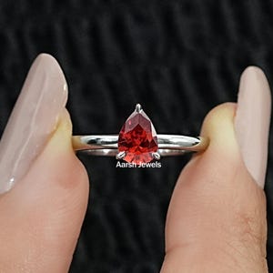 Pode incluir: Um anel de tom prateado com uma gema vermelha de corte em forma de pera num engaste de garras. A banda é lisa e polida. O texto Aarsh Jewels está impresso em letras brancas pequenas logo abaixo da gema vermelha na banda do anel.
