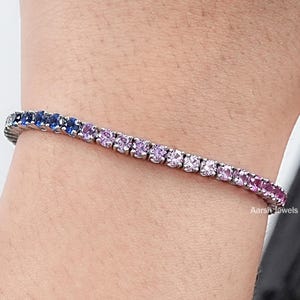 May include: A tennis bracelet with a gradient of colorful gemstones. The bracelet transitions from dark blue to purple, pink, and red. The bracelet is worn on a wrist.