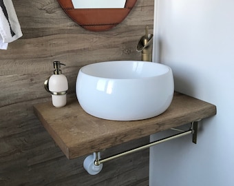 May include: A white ceramic vessel sink with a gold tap mounted on a wooden worktop. The worktop is attached to the wall with a gold metal bracket and a towel rail. The background features a brown wood-look wall with a brown leather-wrapped mirror.