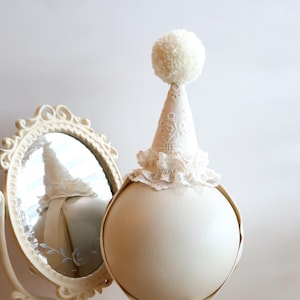 May include: A cream-coloured spherical display stand featuring a pointed hat topper with lace trim and a white pom-pom. The stand includes a ribbon tie with two small tassels at the ends. Beside it sits an ornate, oval-shaped cream vanity mirror.