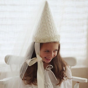 May include: A young girl wearing a white quilted cone-shaped hat with a lace trim and a white veil. She is wearing a white dress with lace detailing and a white ribbon tied around her neck. She is smiling and looking down.