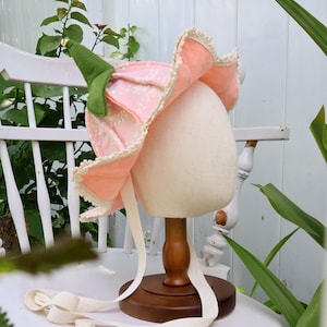 May include: A bonnet shaped like a flower with light pink petals featuring a white floral pattern, a green stem-like top with a small gold bell, and cream-colored lace trim along the edges. The bonnet includes long cream-colored fabric ties.