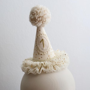 May include: A cream-colored birthday party hat with a gold embroidered number 1, gold star accents, and a fluffy pom-pom. The hat has a ruffled lace trim and is displayed on a cream-colored sphere.