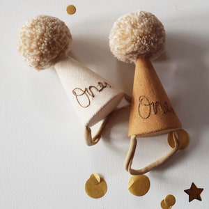 Op de afbeelding: Twee beige feestmutsen met pompoenen erop. De mutsen zijn versierd met het woord "One" in bruine borduurwerk. De mutsen liggen op een witte achtergrond met gouden confetti.