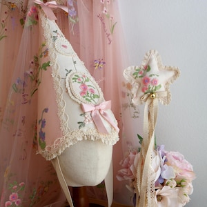 May include: A cone-shaped hat made of cream fabric with floral embroidery, lace trim, and a pink satin bow, paired with a star-shaped wand featuring floral embroidery, a small bell, and long lace ribbons, set against a sheer pink floral cape.