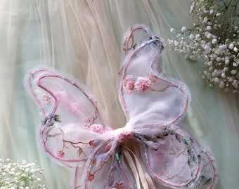 May include: Delicate white butterfly wings with pink floral embroidery and a cream-colored ribbon tie. The wings are on a light green and cream-colored fabric background.
