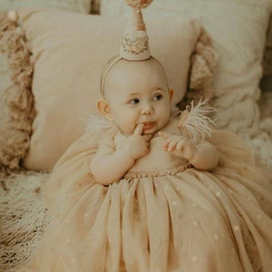 K&ouml;nnte beinhalten: Ein kleines M&auml;dchen in einem beigefarbenen T&uuml;llkleid mit Blumenmuster und einer wei&szlig;en Partym&uuml;tze mit einem rosa Bommel. Das Baby sitzt auf einer wei&szlig;en, flauschigen Decke und schaut nach rechts.