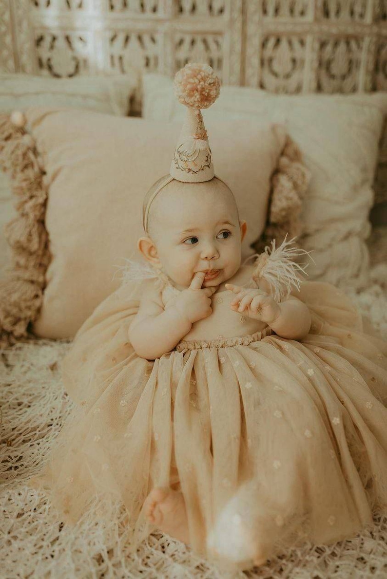 K&ouml;nnte beinhalten: Ein kleines M&auml;dchen in einem beigefarbenen T&uuml;llkleid mit Blumenmuster und einer wei&szlig;en Partym&uuml;tze mit einem rosa Bommel. Das Baby sitzt auf einer wei&szlig;en, flauschigen Decke und schaut nach rechts.