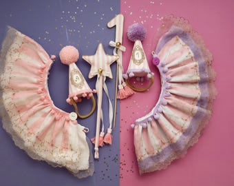 May include: A collection of party accessories in pink, white, and gold. Includes a star wand, a number 1, two party hats, and two ruffled collars. The items are decorated with stripes, stars, and pom-poms, perfect for a first birthday celebration.