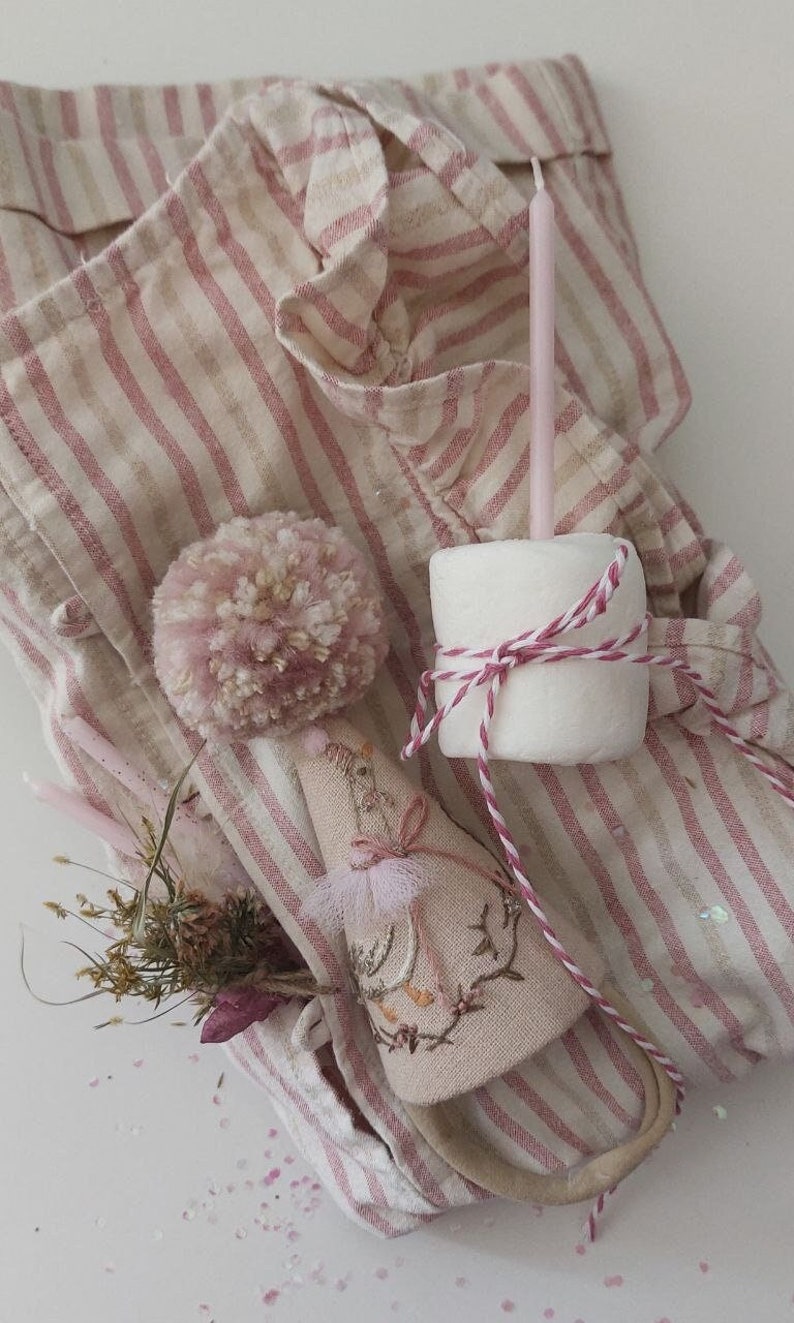 K&ouml;nnte beinhalten: Ein rosa-wei&szlig; gestreiftes Stoff mit einem rosa Bommel, einem kleinen gestickten Vogel und einem wei&szlig;en Marshmallow mit rosa-wei&szlig;em Garn. Es gibt auch rosa-wei&szlig;e Kerzen und rosa Glitzer auf dem Stoff.