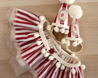 May include: A set of red and white striped party accessories, including a ruffled collar with lace trim and two cone-shaped hats. The hats have white pom-poms and are decorated with the letter "A" and the number "1/2".