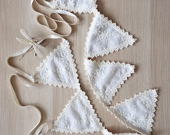 May include: White lace bunting with floral embroidery and scalloped edges. The triangular flags are connected by a cream-colored ribbon. A decorative dragonfly and tassel are also included.