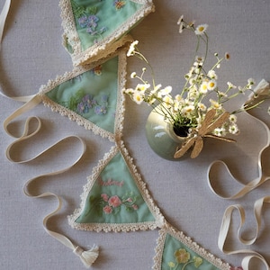May include: A mint green fabric banner with embroidered floral designs and cream-colored lace trim. The banner is strung with a cream-colored ribbon and has a tassel at the end. A small vase with flowers and a dragonfly decoration is also present.