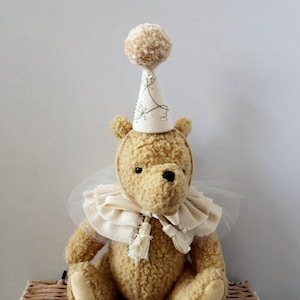 Peut inclure: Un ours en peluche brun clair portant un chapeau de fête avec un pompon et un col à volants. L'ours a un chapeau et un col de couleur crème. L'ours est assis sur un panier tressé.