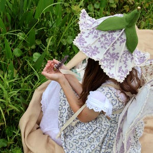 Pode incluir: Uma criança com um chapéu em forma de flor e asas de fada sentada na grama. O chapéu é branco com um padrão floral roxo e folhas verdes. O vestido é branco com um padrão floral azul. A criança segura um raminho de lavanda.