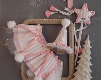 May include: A collection of festive party decorations in shades of pink and white. Includes a striped number one, a star-shaped wand, a party hat, a decorative tree, and a garland with pom-poms. The items are arranged on a gray background.